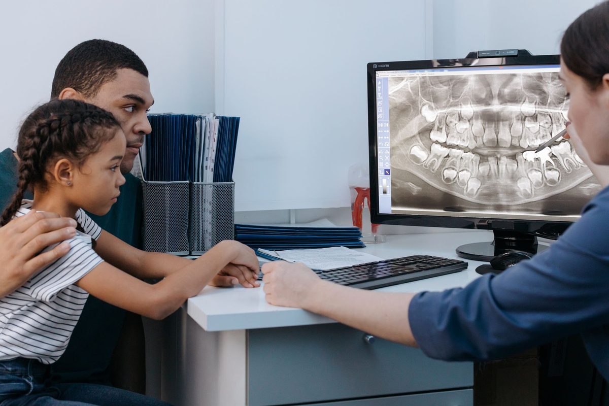 A father and his young daughter looking at a dental X-ray on a monitor with a clinic staff member
